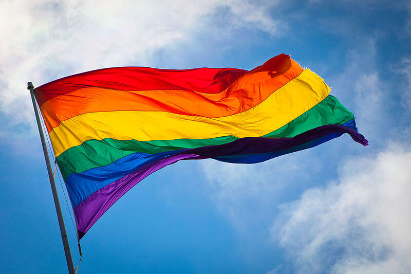 The rainbow-striped queer pride flag flying in front of a partially cloudy sky background